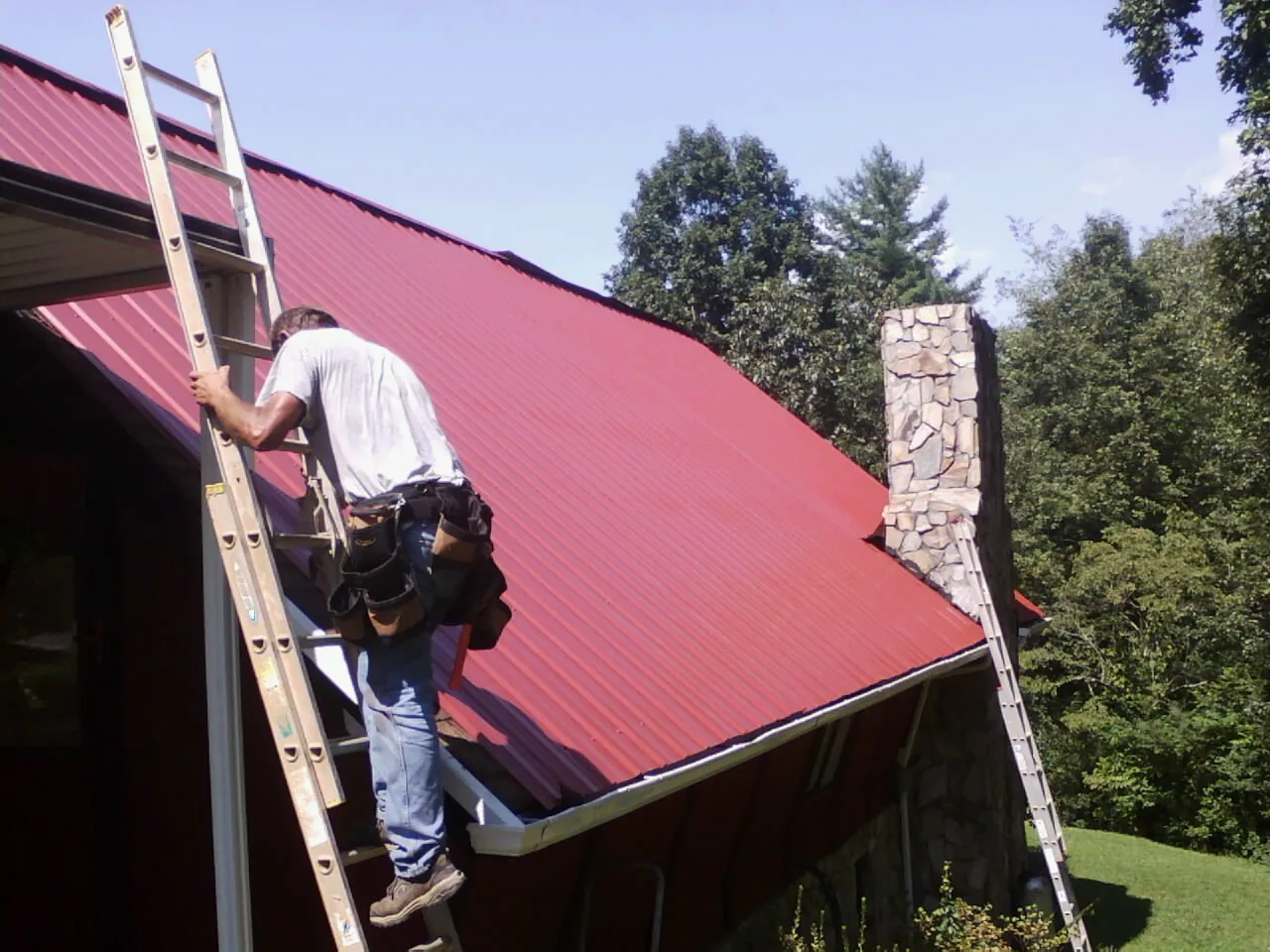 Roofer on a ladder beside a red metal roof and stone chimney — Soffit & Fascia Repair by Roofing Brothers in Seneca